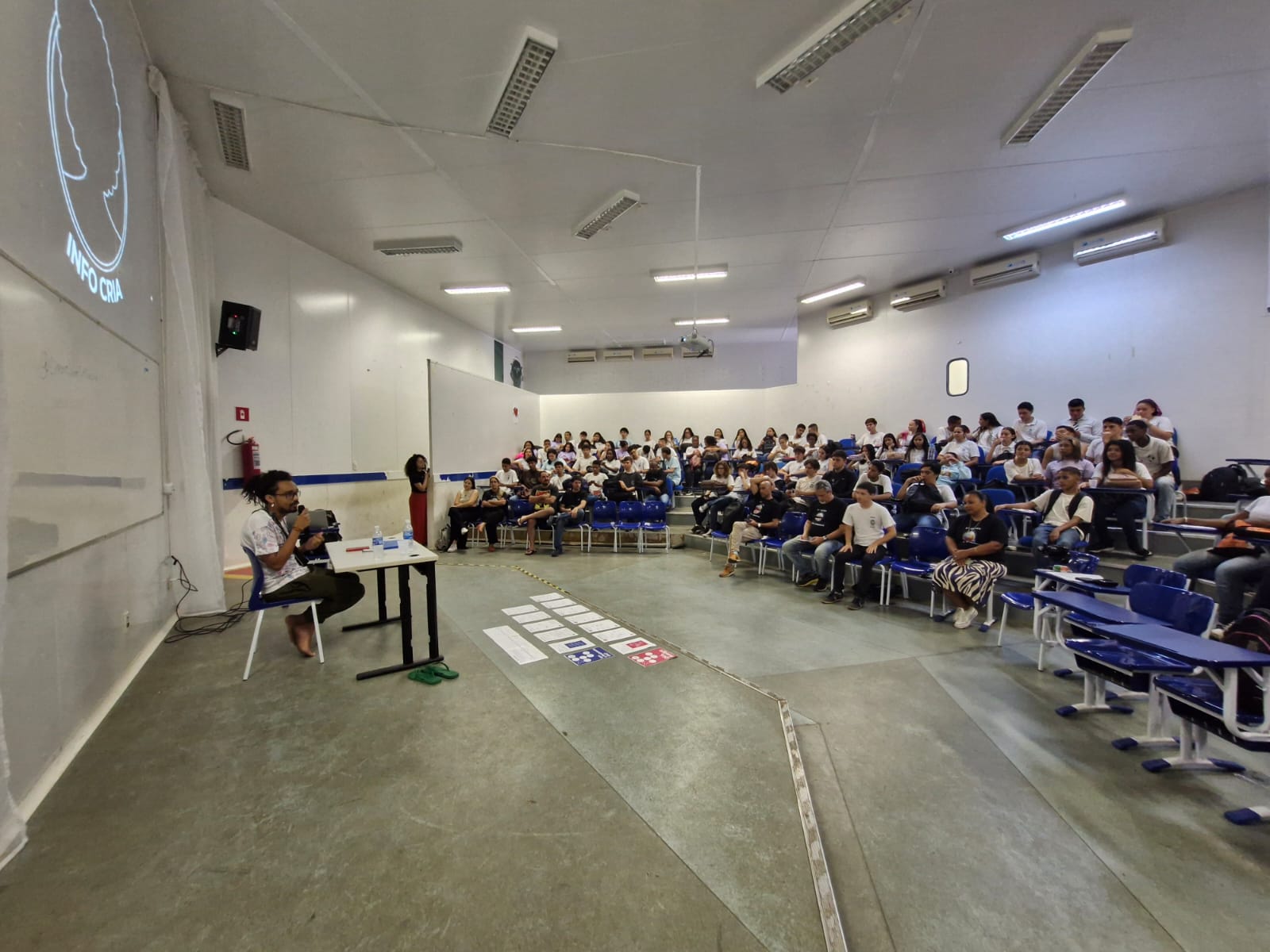 vista do auditório da FAETEC de Imbariê durante a oficina, com a mediação à esquerda e participantes nas cadeiras.