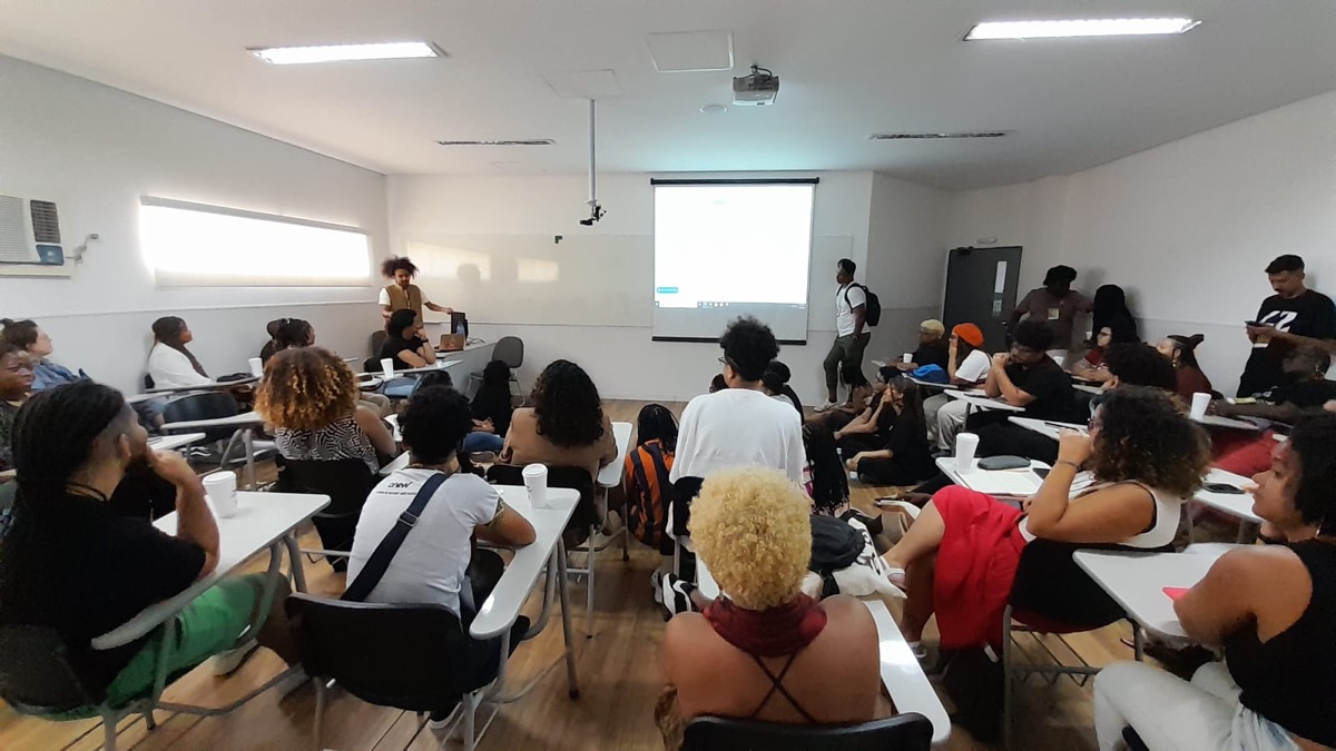 Raffael à frente de uma sala durante uma palestra, com participantes sentados e uma projeção ao fundo.